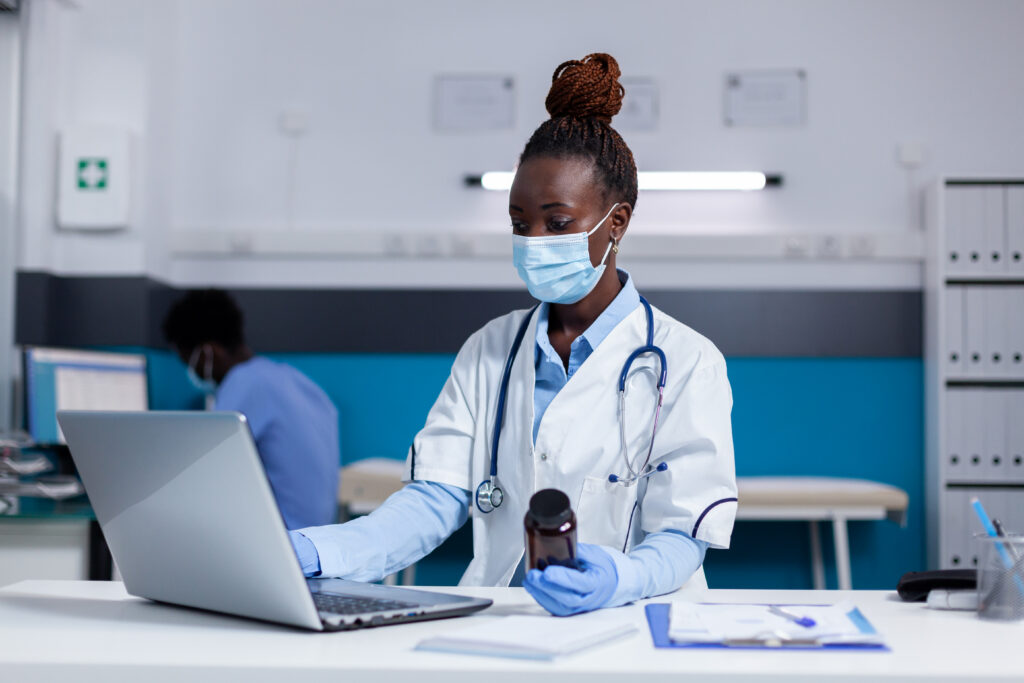 African american woman with doctor job holding bottle of medicine while using laptop on white desk in medical office. Black medic with uniform and stethoscope looking at digital screen